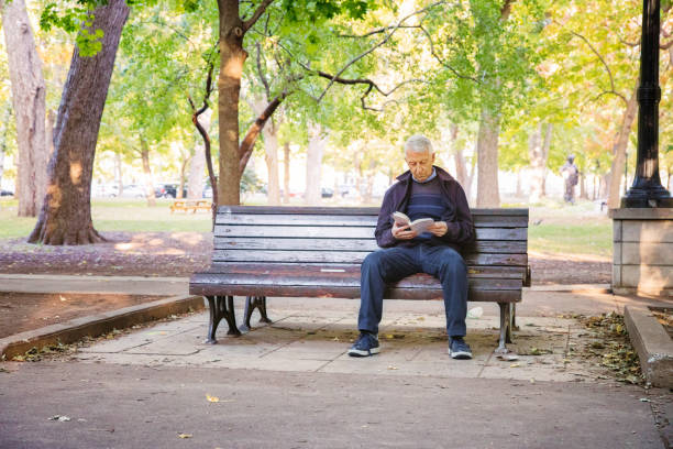 uomo anziano che legge il libro da solo nel parco pubblico - men reading outdoors book foto e immagini stock