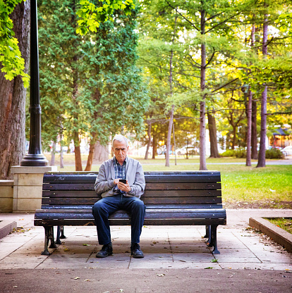 Senior man watching media on mobile phone alone in a public park on a beautiful Autumn afternoon. He is frowning.