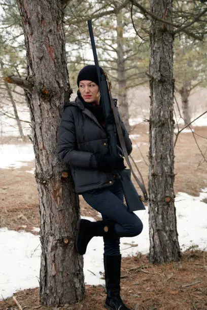 Photo of young woman in winter forest with shotgun stands near a tree