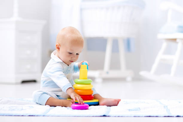 Baby playing with toy pyramid. Kids play Cute baby playing with colorful rainbow toy pyramid sitting on play mat in white sunny bedroom. Toys for little kids. Interior for little boy nursery. Child with educational toy. Early development. baby toys stock pictures, royalty-free photos & images