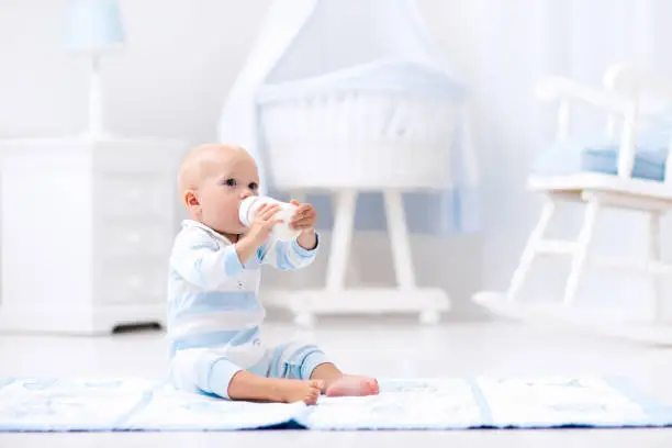 Adorable baby boy playing on a blue floor mat and drinking milk from a bottle in a white sunny nursery with rocking chair and bassinet. Bedroom interior with infant crib. Formula drink for infant.