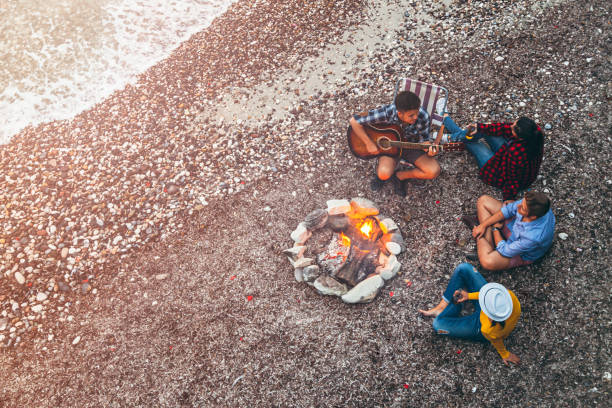 Young friends enjoying music near campfire Aerial view of best young friends having fun near the bonfire while camping on beach. Drinking beer, playing guitar and listening music against sea and sky background. Bonfire stock pictures, royalty-free photos & images