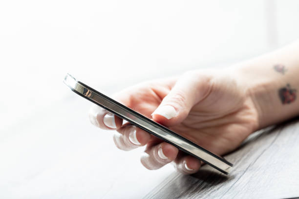 Young woman with tattoos on her wrist holding a modern smartphone in her hand as she texts a message, close up side view