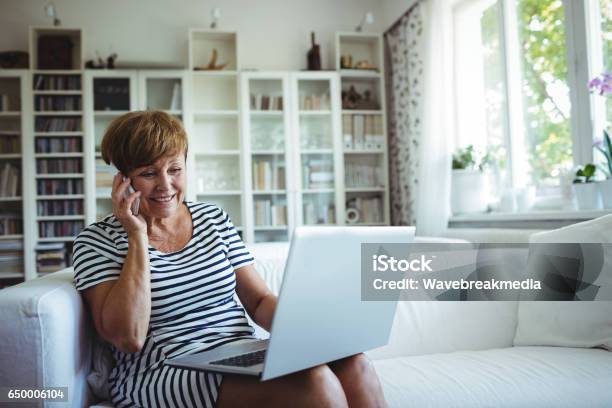 Ältere Frau Am Handy Während Mit Laptop Im Wohnzimmer Stockfoto und mehr Bilder von Alter Erwachsener