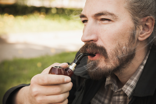 Young serious asian bearded man smoking pipe in summer park, close up portrait