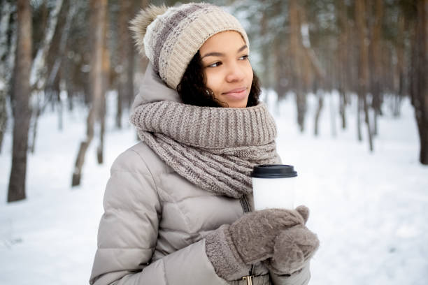 Dreamful young woman standing in winter forest and looking aside. Pensive African woman with take-away coffee contemplating nature around. Park concept
