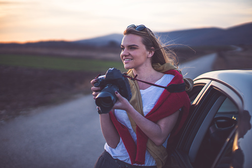 Young woman standing next to the car on the road and taking phoos with digital camera of a beautiful sunset in the spring