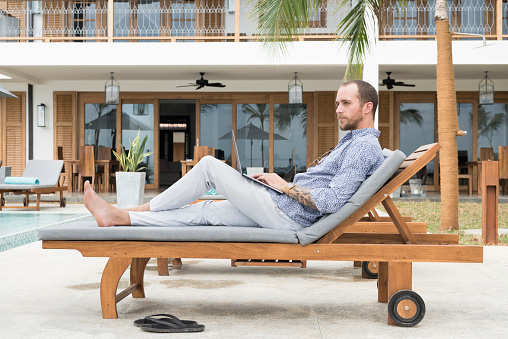 Man in his 30s reclining on seat at poolside, working remotely, luxury villa in background, businessman on vacation in Sri Lanka