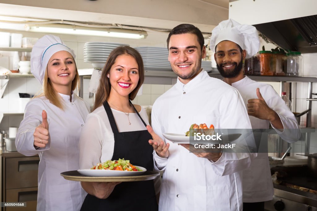 Portrait of positive kitchen workers Happy chefs and young smiling nippy in apron working at restaurant kitchen. Selective focus Adult Stock Photo