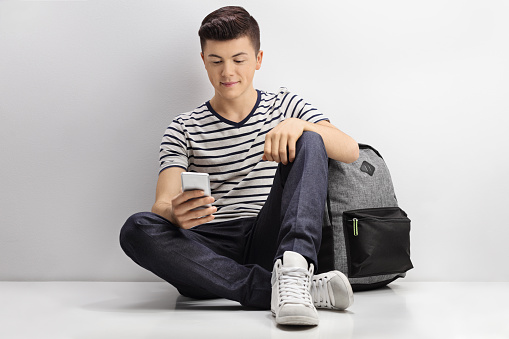 Teenage boy with a backpack looking at a phone and leaning against a gray wall
