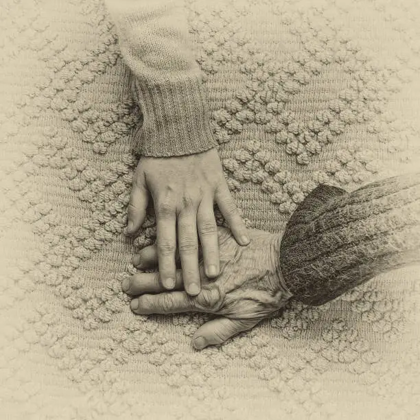Granddaughter gently holding her hand over her grandmother’s hand. Hands are lying on a picturesque blanket. Close up. Selective focus. Post processed to look like an old photo.