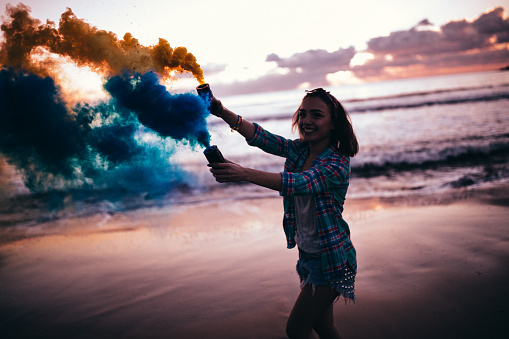 Beautiful young hipster woman playing with colorful smoke bombs at the beach after sunset