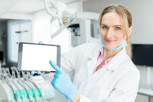 Horizontal color image of female dentist showing  blank screen on digital tablet in dental clinic office, smiling and looking at camera. Female doctor wearing white uniform, blue surgical mask and gloves. Dental equipment in the background.