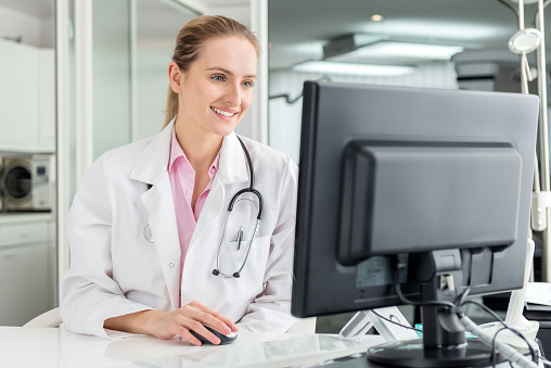 Horizontal color portrait of pretty female doctor at office sincerely smiling and looking at a computer. Wearing white lab coat and stethoscope around neck.