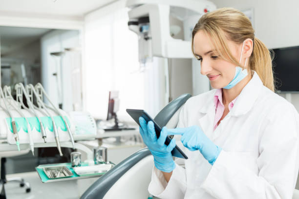 Female dentist touching tablet at the clinic Horizontal color image of female dentist touching digital tablet in dental clinic office, sitting and smiling. Female doctor wearing white uniform, blue surgical mask and gloves. Dental equipment in the background. dental drill stock pictures, royalty-free photos & images