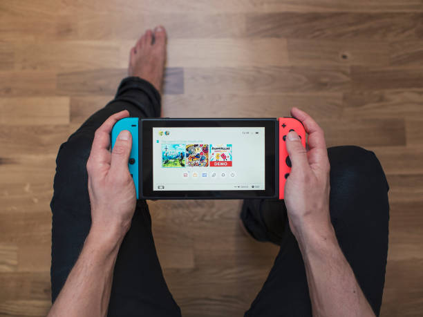 Nintendo Switch neon Game Console Gothenburg, Sweden - March 10, 2017: A shot from above of a young man's hands holding a neon coloured Nintendo Switch video game system developed and released by Nintendo Co., Ltd. in 2017. The system is turned on and its main menu is showing on the display. Shot on a hardwood floor background in a home environment. brand name games console stock pictures, royalty-free photos & images