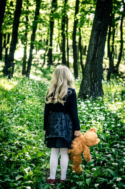 lost girl in black dress holding teddy bear toy in the spring forest back view