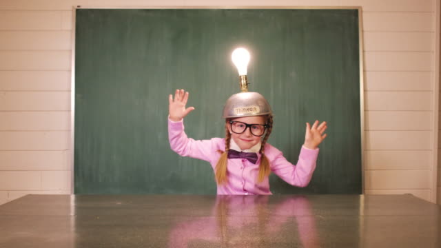 A young girl dressed as a nerd with a homemade thinking cap intensely searches for that new big idea. The girl with the light bulb helmet succeeds and the light comes. She celebrates success with arm gestures and a crazy face.
