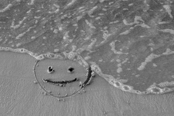 On the sand drawn a smiley face and a wave washes, black and white stock photo