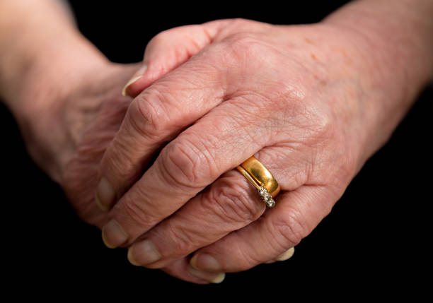 Close up of an elderly married lady's hands clasped in front of her in a relaxed position. On a black background.