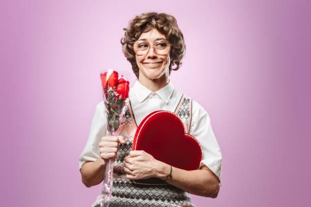 Adult Nerd Woman Looking for Love A woman in 1980's style and fashion wears a heart covered sweater vest while posing in front of a pink background. embarrassed-smile stock pictures, royalty-free photos & images