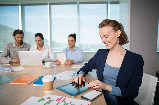 Smiling female business executive using digital tablet in conference room while colleagues working in background