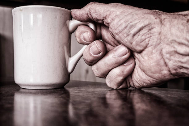 Closeup of unrecognizable aged person with wrinkles and veins holding mug of hot coffee in hand against blurred interior in light