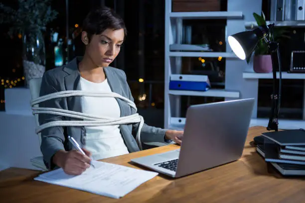 Businesswoman tied with rope while working on laptop at her desk in office at night