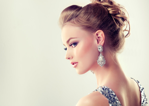 Portrait of young gorgeous girl dressed in evening gown,with delicate makeup on her face and large jewelry earrings on ears. Example of evening or wedding hairdress.