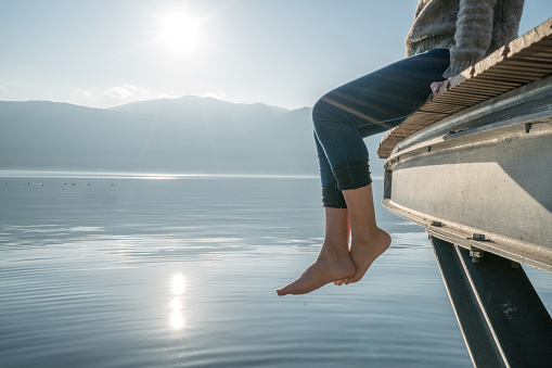 Woman's feet dangle from wooden wharf, above lake. Shot in Italy, sunbeam, reflection on water surface.