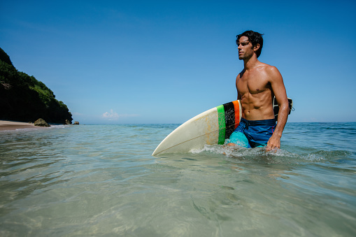 Shot of young man with surf board walking out of the sea. Handsome caucasian male holding surfboard coming out of the ocean.