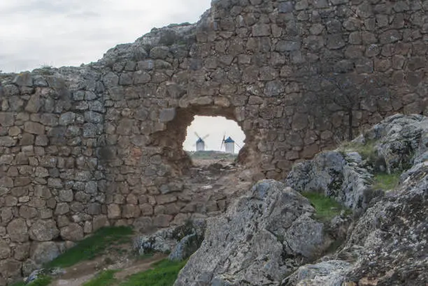 Photo of White old windmills on the hill near Consuegra (Castilla La Mancha, Spain), a symbol of region and journeys of Don Quixote (Alonso Quijano) and a town on cloudy day, a view through a hole in the wall.