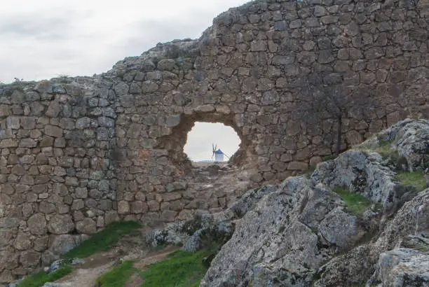 Photo of White old windmill on the hill near Consuegra (Castilla La Mancha, Spain), a symbol of region and journeys of Don Quixote (Alonso Quijano) and a town on cloudy day, a view through a hole in the wall.