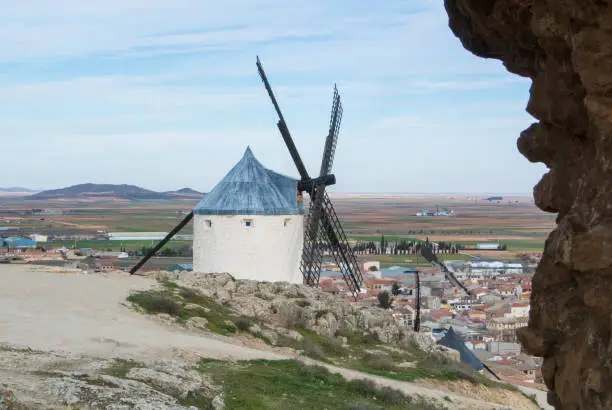 Photo of White old windmills on the hill near Consuegra (Castilla La Mancha, Spain), a symbol of region and journeys of Don Quixote (Alonso Quijano) and a town on cloudy day.