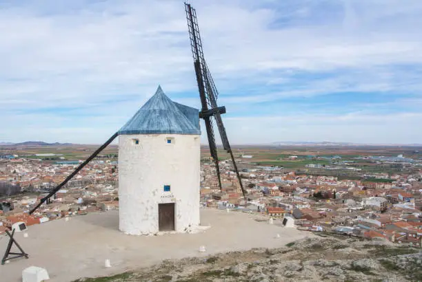 Photo of White old windmill on the hill near Consuegra (Castilla La Mancha, Spain), a symbol of region and journeys of Don Quixote (Alonso Quijano) and a town on cloudy day.