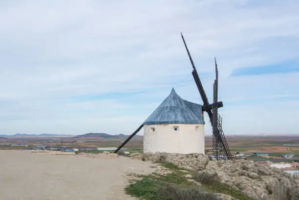 Photo of White old windmill on the hill near Consuegra (Castilla La Mancha, Spain), a symbol of region and journeys of Don Quixote (Alonso Quijano) and a town on cloudy day.