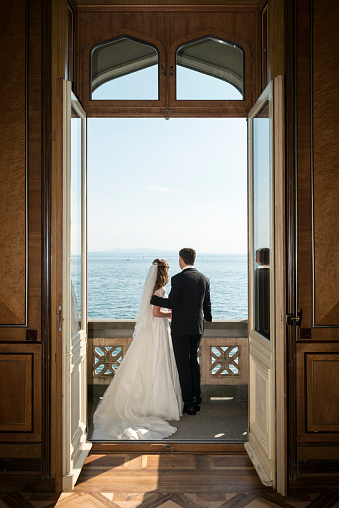 View through open doors at a happy smiling bridal pair standing at the balcony of an old villa with a lake in the background. The bride is wearing a beautiful white dress, the groom is wearing a elegant black suite.