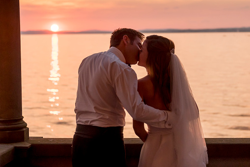 Happy bridal pair kissing at sunset at the balcony of an old villa with a lake in the background. The bride is wearing a beautiful white dress, the groom is wearing a elegant black suite.