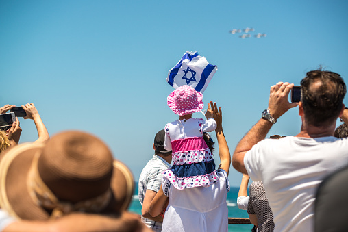 Tel Aviv, Israel - May 12, 2016:  Tel Aviv beaches crowded with people celebrating Independence Day. People photographing with mobile devices air show in Tel Aviv sky.   Little girl holding Israeli flag sitting on mother's shoulders.