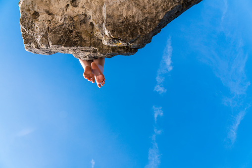 Teenager girl sitting on the edge of the cliff, with her legs in the air against the blue summer's sky. View directly below. Tobermory, Ontario, Canada, North America.