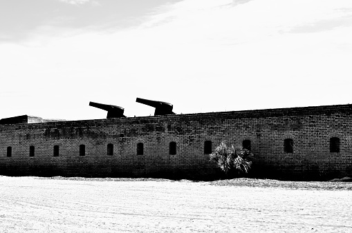 Black and white image of an old historical, military, brick fort on a beach in America, with two cannons pointing to the left atop the brick fort's wall.
