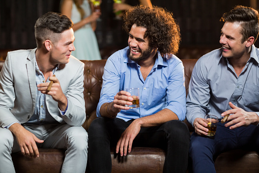 Smiling friends interacting with each other while having cigar and whisky in bar