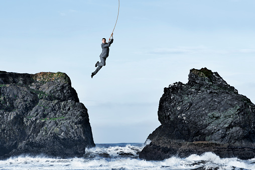 A businessman hangs on for dear life as he swings on a rope from one rocky cliff to another as the ocean waves crash against the rocks below him.