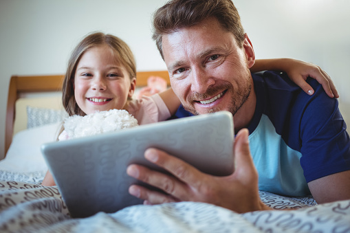 Father lying with daughter on bed and using digital tablet at home