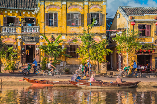 On the river that passes in this beautiful city of Hoi An, we see many boats on the water. Boats are the means of transport for locals and tourists. The boats pass close to the shore and can berth temporarily. On the shores you can see pedestrians and bicycle riders discovering this lovely city teeming with life.