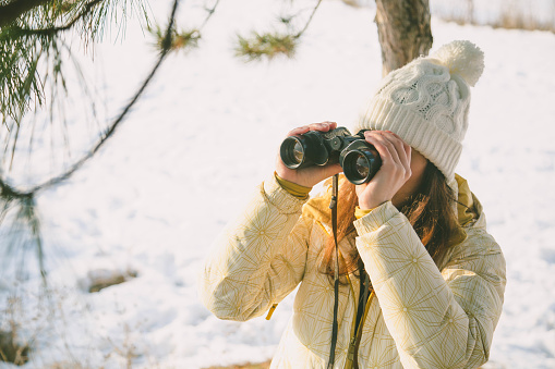 The girl in a White cap with a backpack in the winter in a forest looking through binoculars