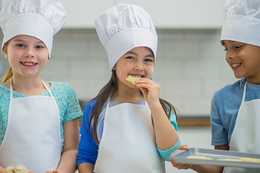 A group of elementary age children are learning how to cook in the kitchen. They are wearing aprons and chefs hats and are testing food.