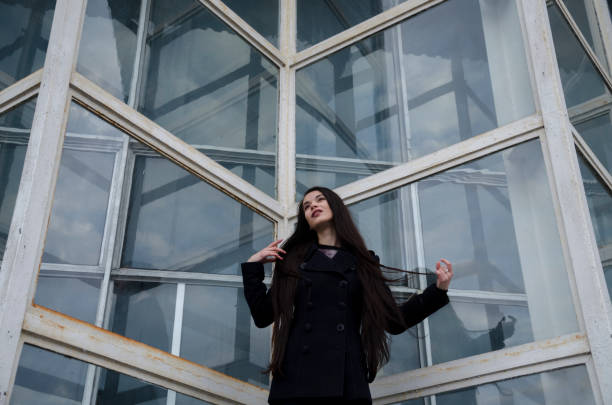 Young beautiful girl with long hair, wearing a hat and wearing a blue coat, on a background of a building made of glass