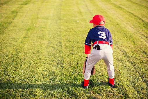 A young Little League baseball player wearing a baseball uniform is waiting for the next play to happen in the outfield. He loves playing America's pastime on a warm summer day in Utah, USA.
