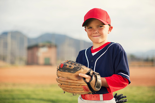 A young Little League baseball player is wearing a baseball uniform and holding his baseball glove while smiling and looking at the camera. He is playing on a warm summer day in Utah, USA.